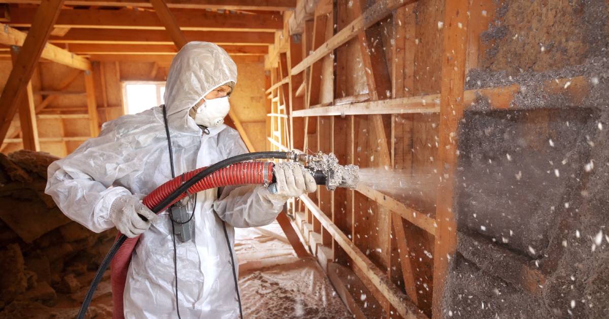 A worker wearing an all-white protective bodysuit and a respirator spraying insulation foam from a hose onto a wall frame.