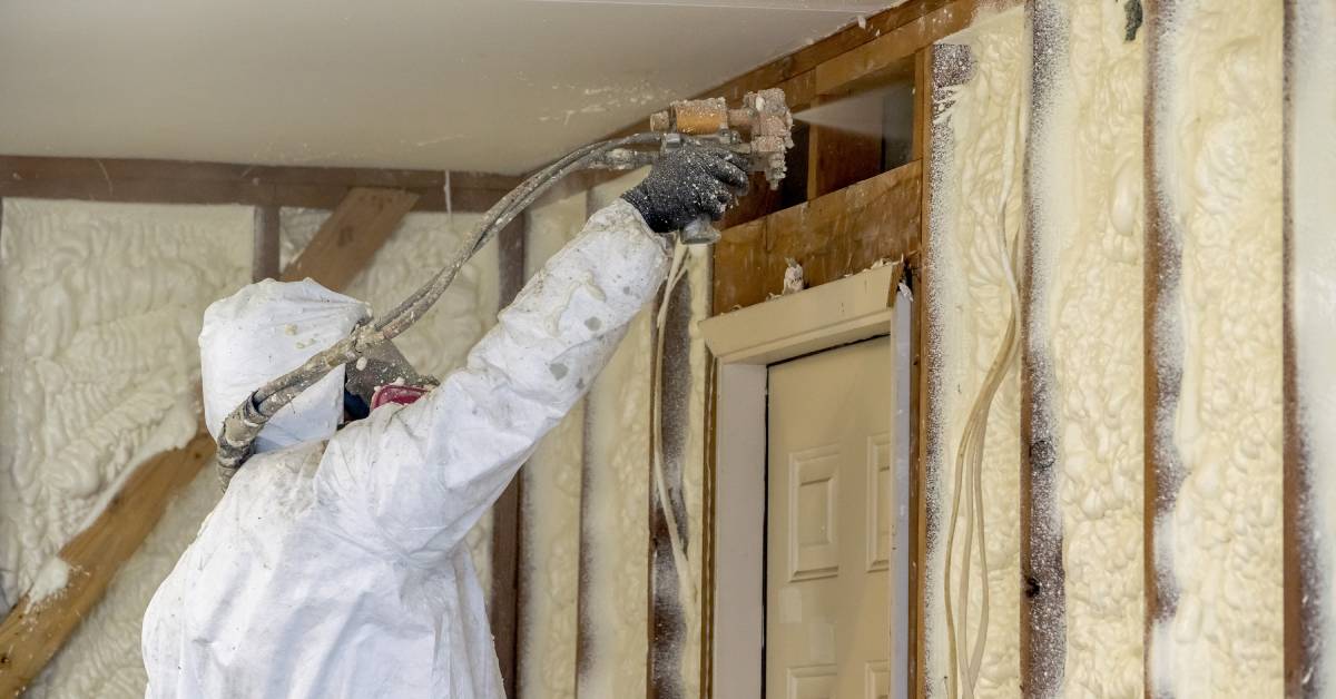 A professional contractor wearing personal protective equipment applies spray foam to the interior walls of a home.