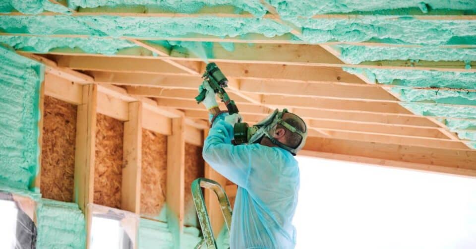 A man wearing PPE, including a respirator and gloves, applying spray foam insulation using a mobile rig.