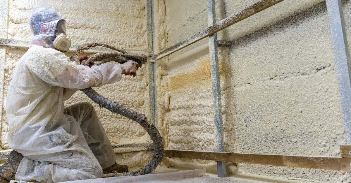A person in protective gear sprays foam insulation onto the walls of a room with a hose inside a construction site.