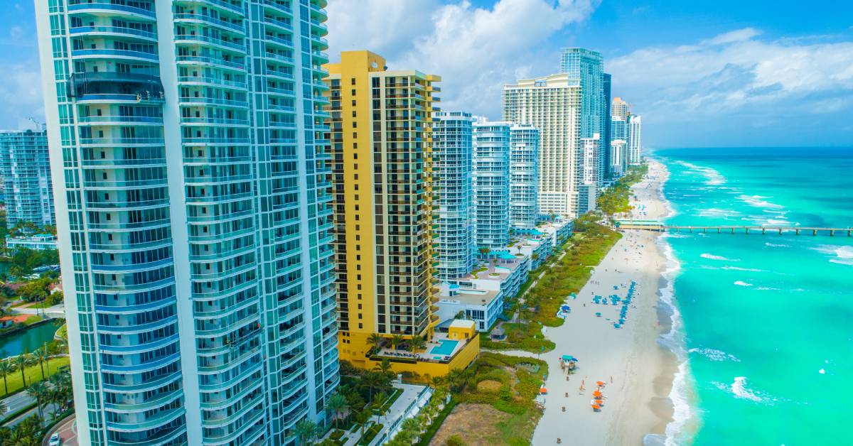 Aerial view of modern coastal high-rise buildings, a beach, and turquoise ocean waves under a partly cloudy sky.