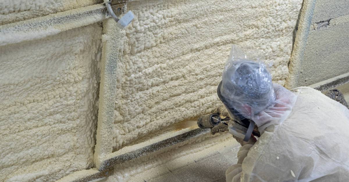 A worker in protective gear sprays foam insulation onto a metal-framed surface, creating a textured insulated layer.