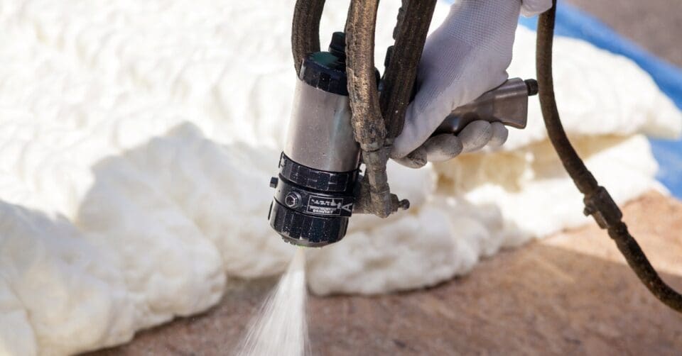 A worker wearing white gloves applies foam insulation to a building's foundation with a precision spray foam gun.