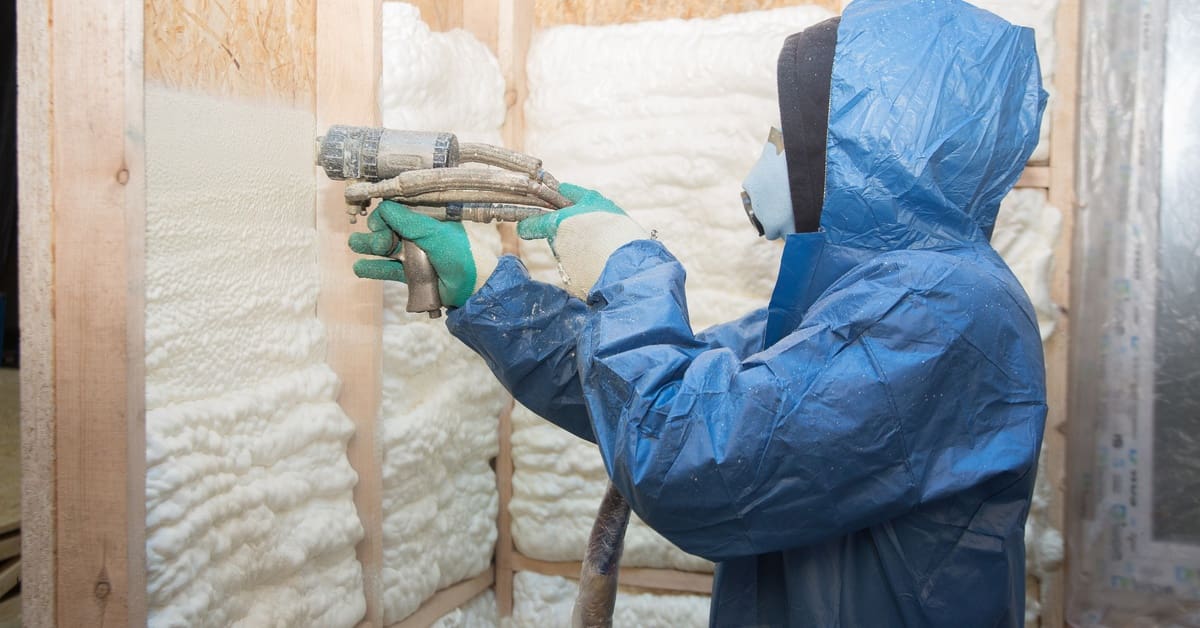 A person in a blue protective suit and gloves spraying white foam insulation on wooden wall frames in an interior space.