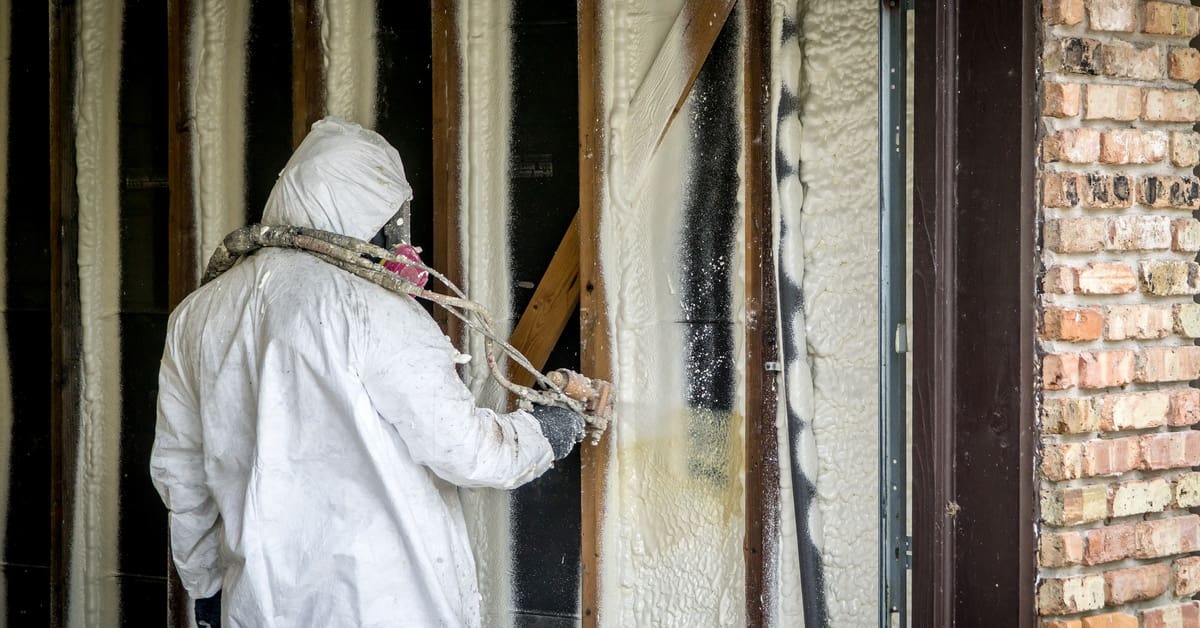 A contractor wearing a white protective suit applies closed-cell spray foam insulation onto a home's walls.