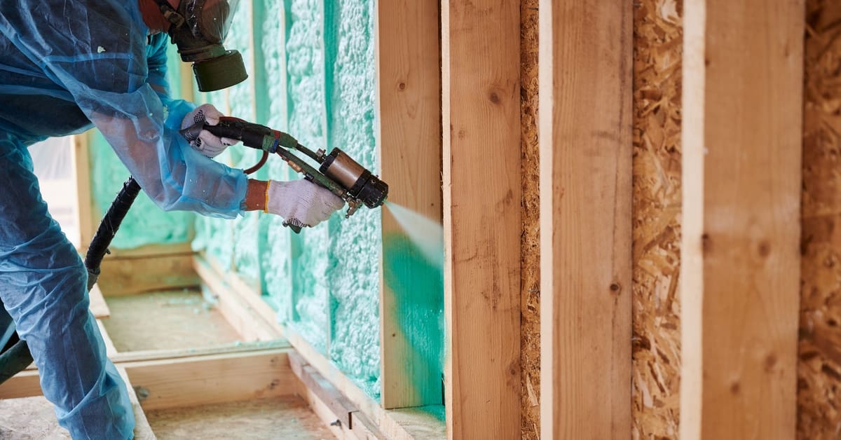 A contractor wearing a blue protective suit and a gas mask sprays insulation onto a home's wooden frames.