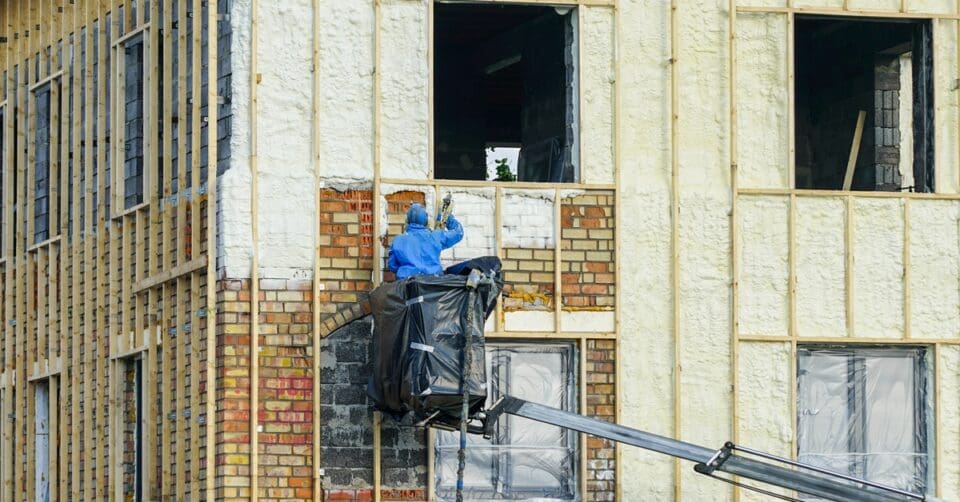 A technician stands on a boom lift and applies spray foam insulation on the building's red brick walls.