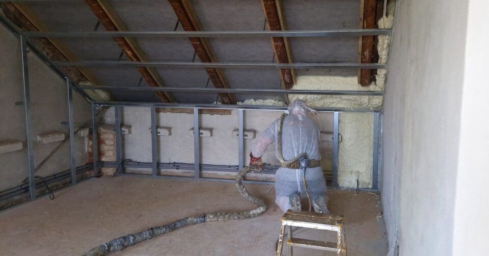 A spray foam technician kneels on the ground as they use a component gun to apply urethane foam to a wall.