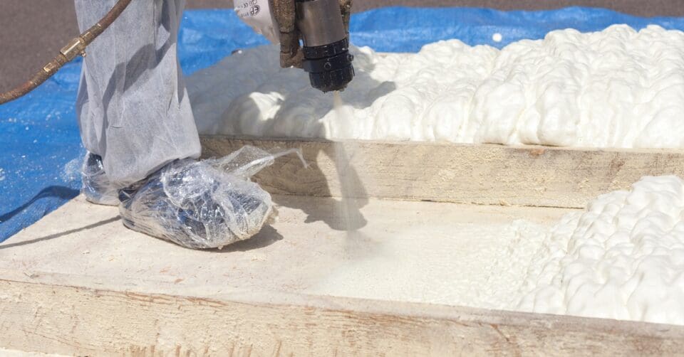 A technician wearing a white protective suit and shoe cover applies spray foam insulation on a wooden frame.