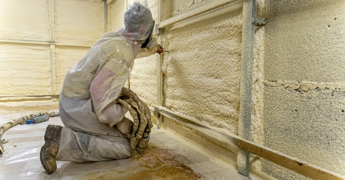 A contractor wearing a protective suit kneels on the ground as they apply polyurethane foam on the walls.
