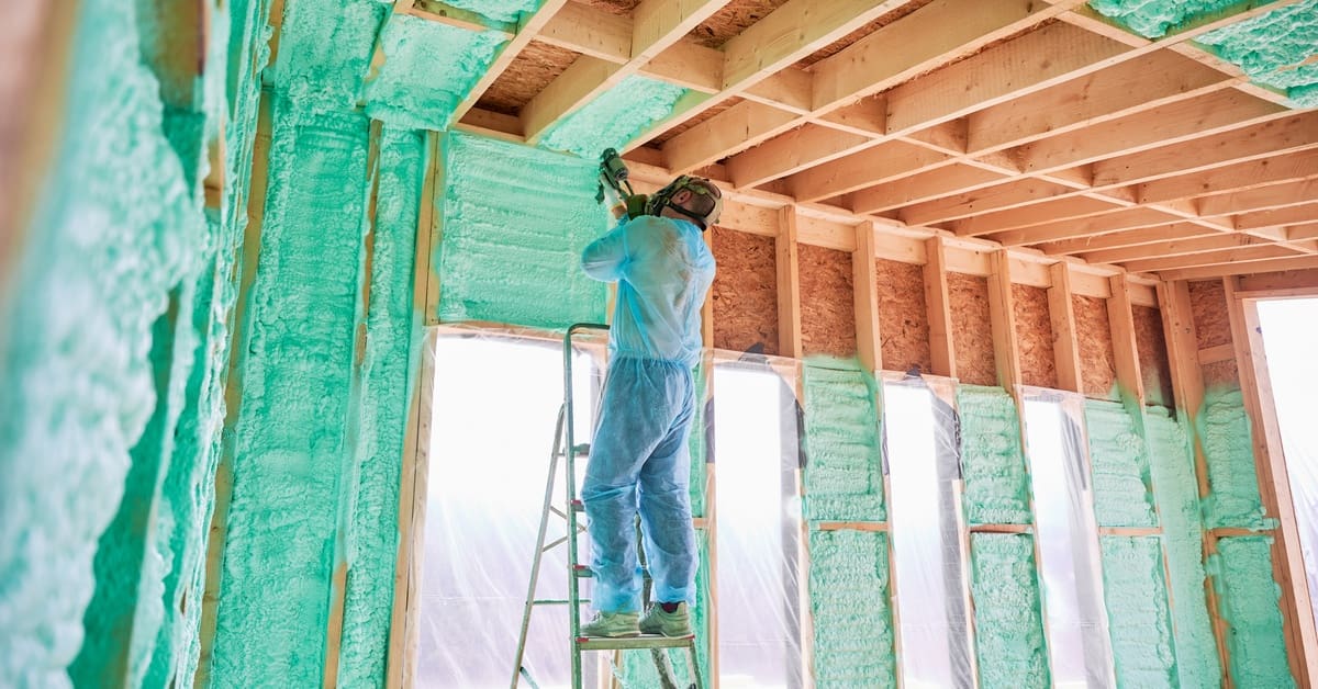 A male builder wearing a gas mask stands on the ladder and applies spray foam insulation to the ceiling.