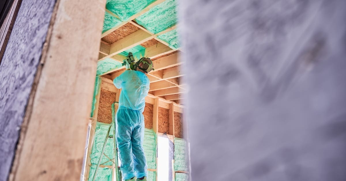 Worker sprays polyurethane foam inside a wood-framed house using a component gun while standing on a ladder.