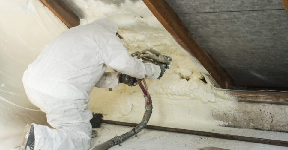 Worker sprays polyurethane foam insulation onto an interior roof surface while wearing protective gear.
