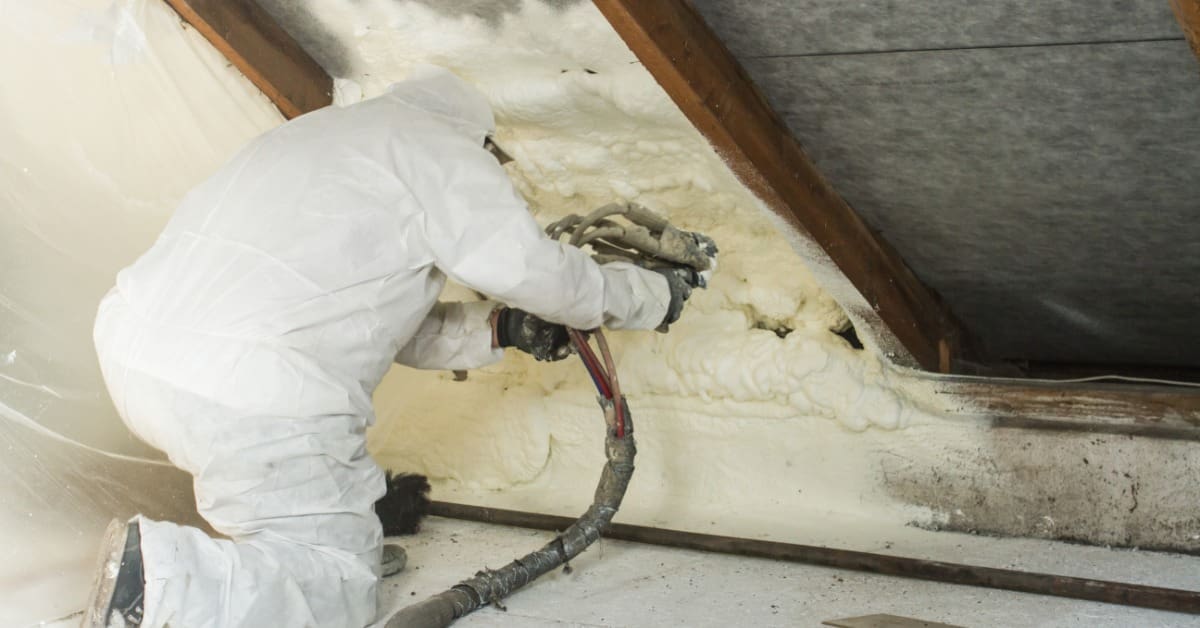 Worker sprays polyurethane foam insulation onto an interior roof surface while wearing protective gear.