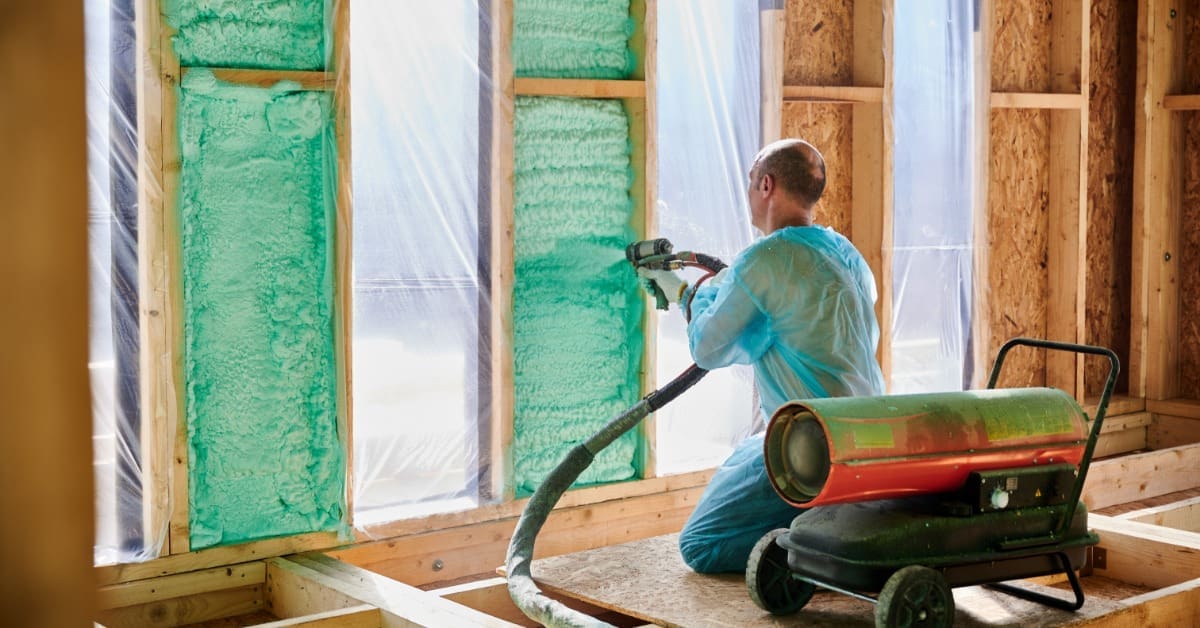a worker spraying foam insulation across a subfloor near wood framing in a partially constructed house interior.