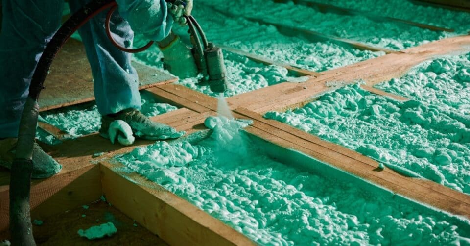 A worker spraying foam insulation between wooden wall studs inside a partially finished residential building.