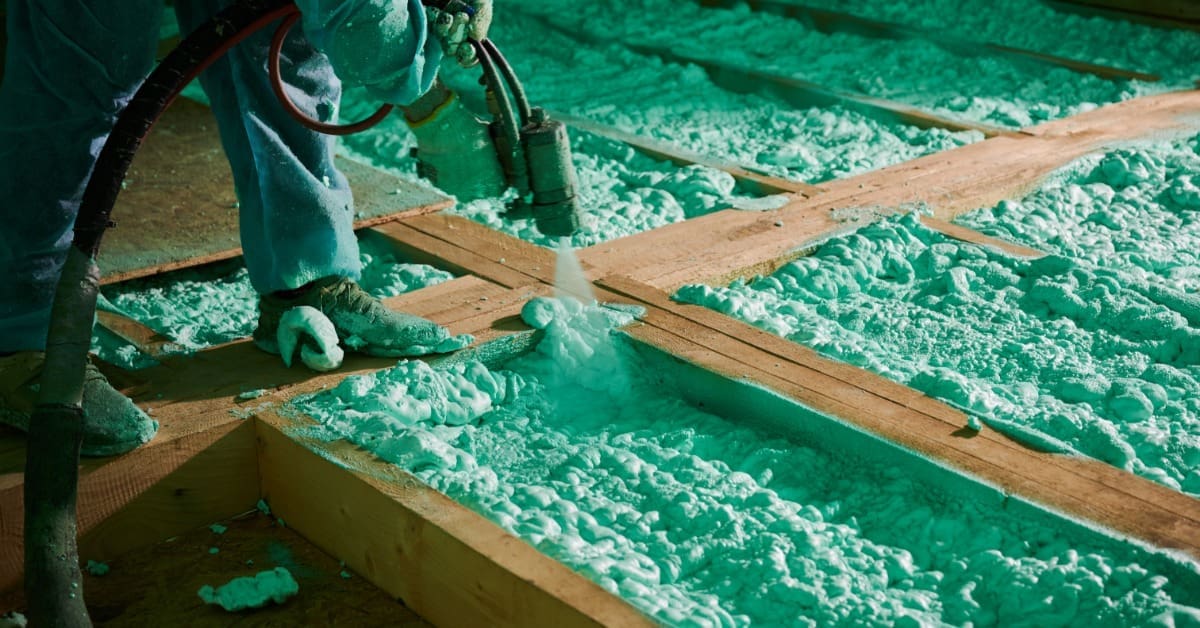 A worker spraying foam insulation between wooden wall studs inside a partially finished residential building.