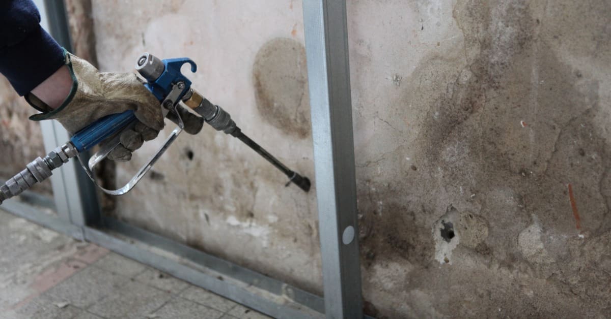 A worker wearing protective gear spraying wall insulation inside a metal-framed residential construction space.