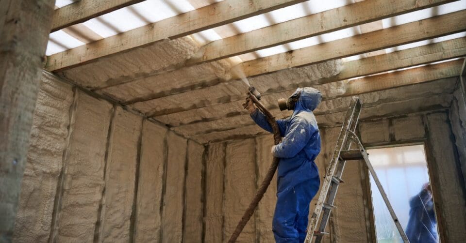 a person in a blue hooded coverall standing beside a ladder while spraying foam insulation onto overhead wooden beams.