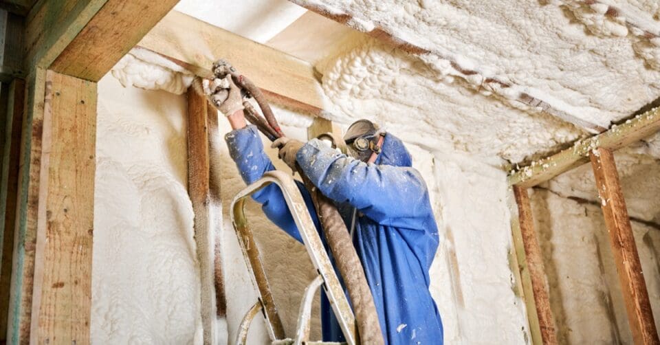 A person in a blue hooded coverall on a ladder spraying foam insulation along wooden framing near a ceiling.