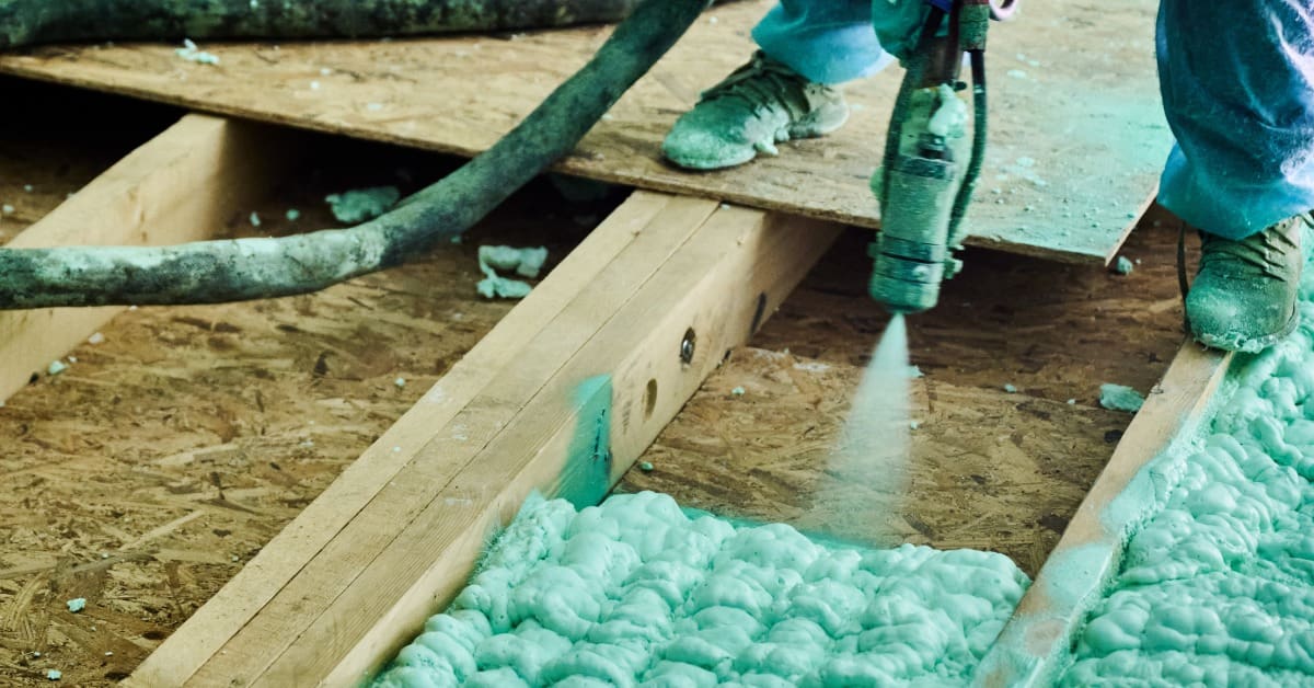A worker spraying pale green foam insulation onto a wooden floor area between exposed beams using a hose and spray gun.