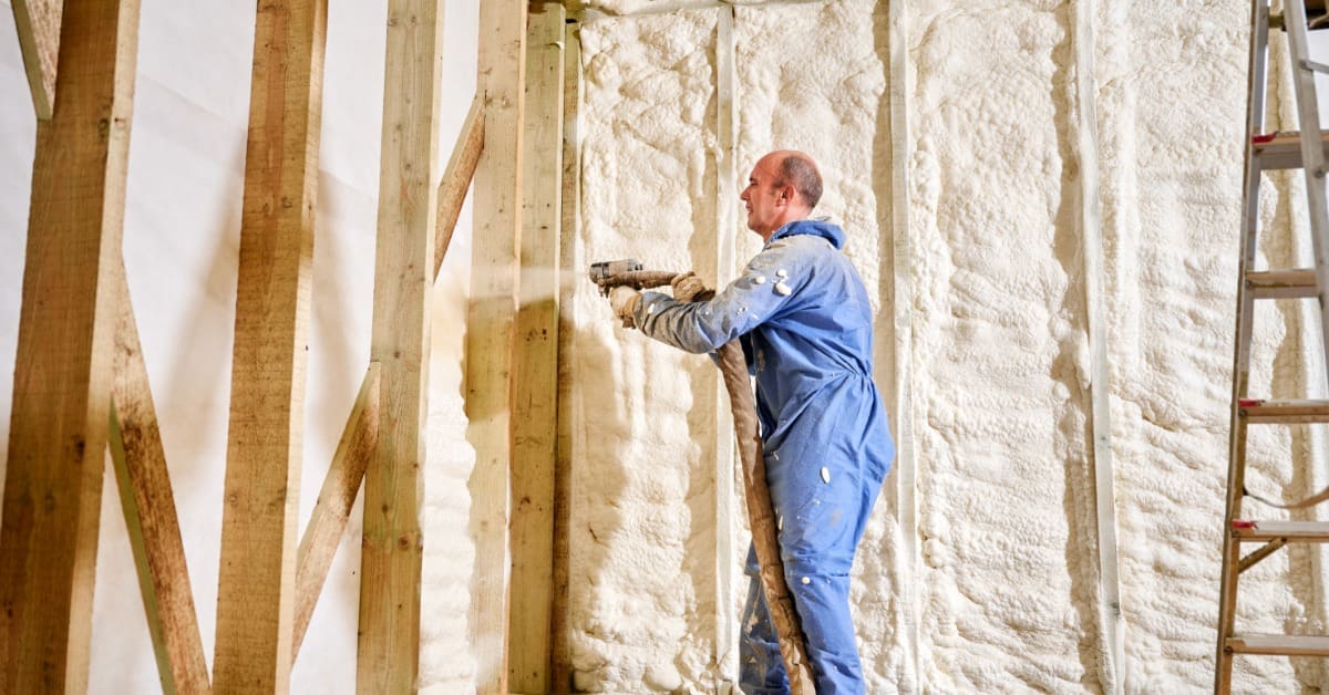 A worker in blue coveralls spraying white foam insulation between wooden wall studs in an unfinished room.
