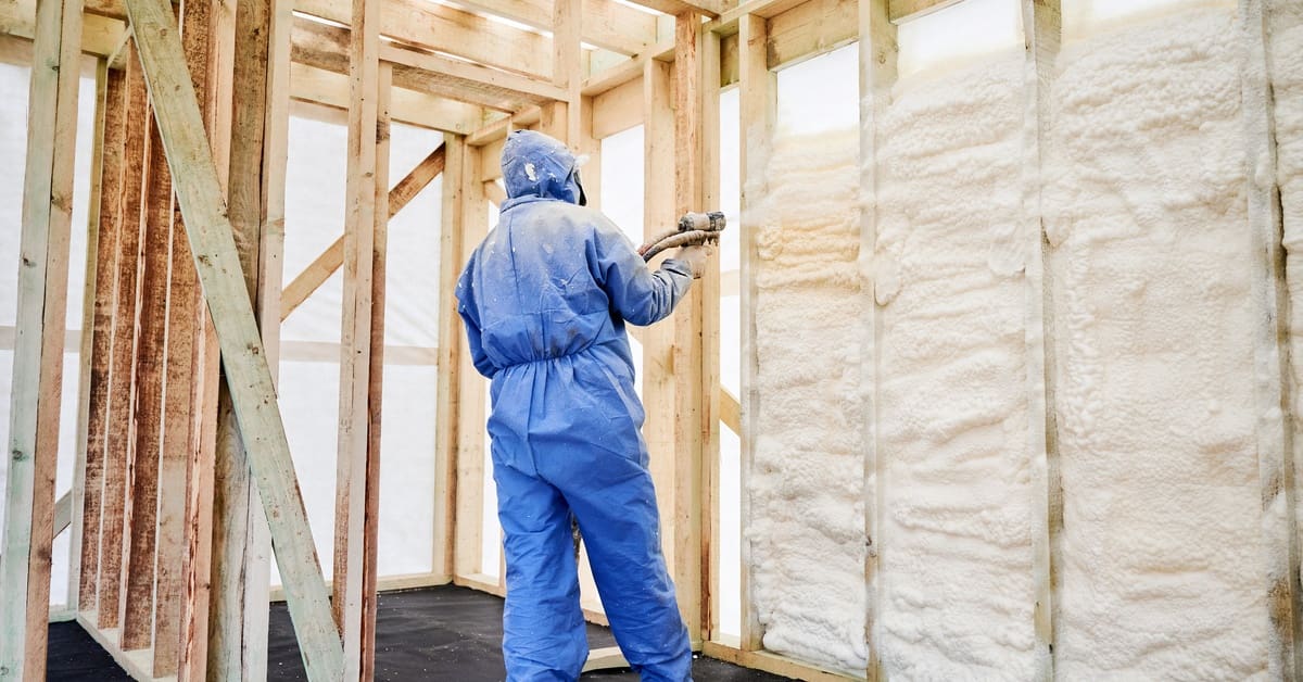 A person in a blue protective suit spraying white foam into an unfinished wood wall with open cavities.