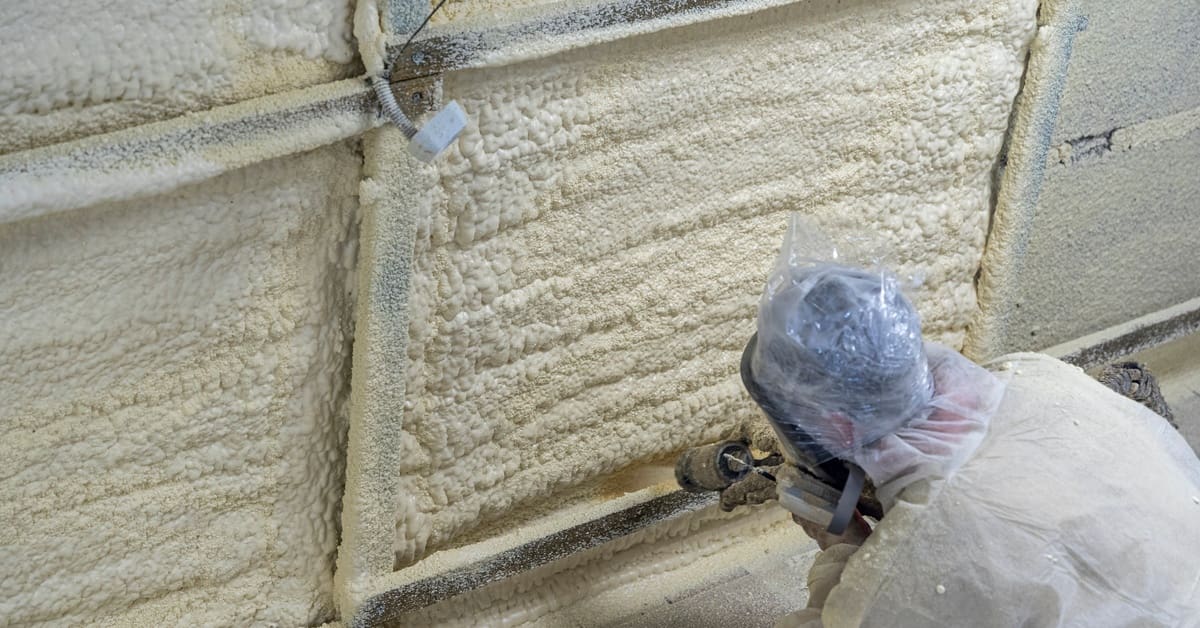 A person in a protective suit kneeling beside a wall while spraying foam insulation with a hose and nozzle.