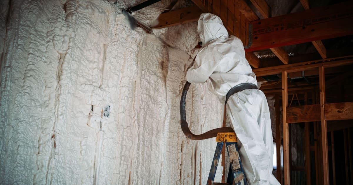 A person in a white protective suit stands on a ladder, spraying foam insulation across exposed interior walls.