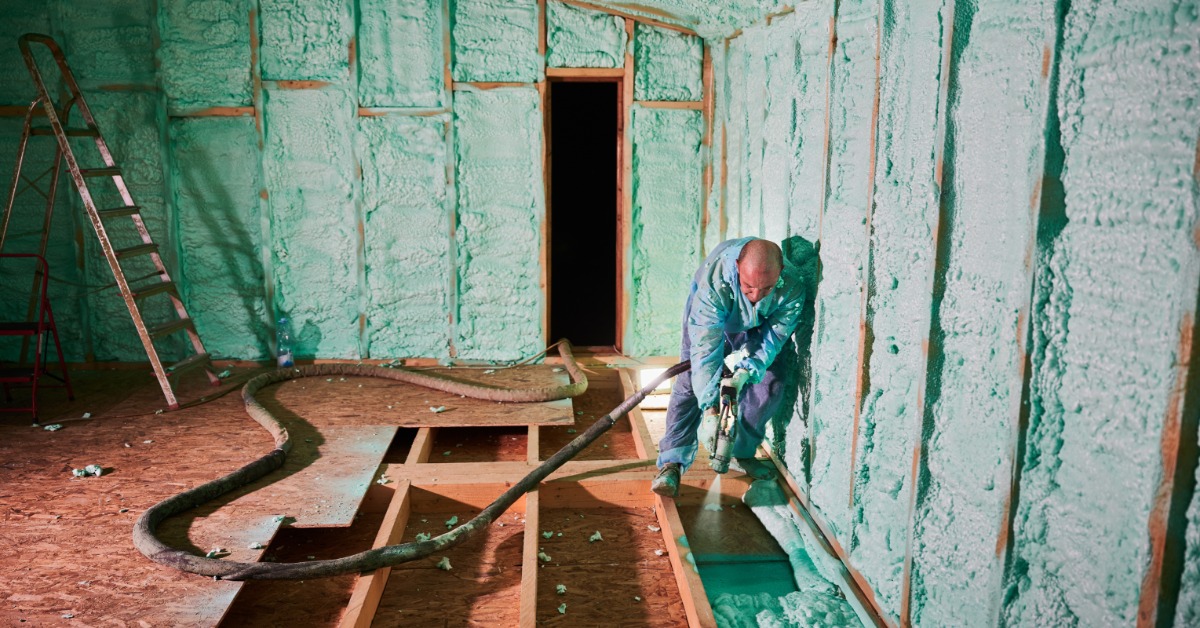 A man spraying green foam insulation along wooden walls inside an unfinished room with a ladder and hoses nearby.
