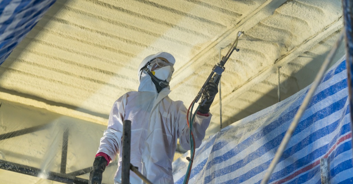 A person in a white protective suit spraying foam insulation onto a ceiling beside striped plastic sheeting.