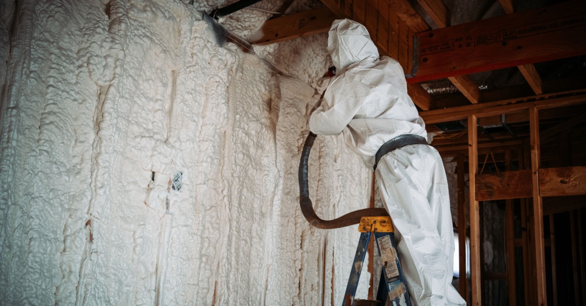 A person in a white protective suit standing on a ladder and spraying foam insulation onto unfinished walls.